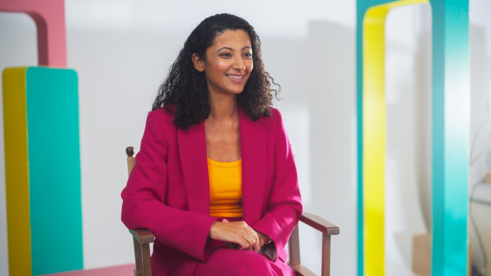 Young Female Host In Vibrant Pink Suit And Yellow Top, Smiling Engagingly On Colorful Set, Exemplifies Modern Videography And Dynamic Storytelling In Media Production.