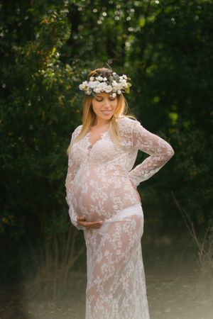 Pregnant woman with closed eyes in white lace dress touching chin while standing in forest