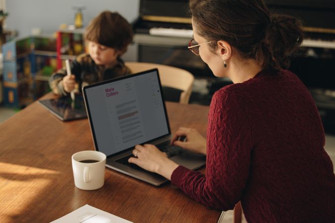 Female parent working from home with laptop and also watching her son