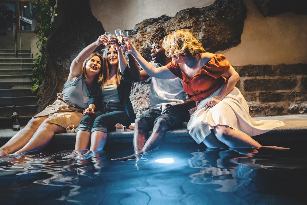 A group of multi-ethnic young people toast by clinking champagne flutes sitting inside the luxury spa resort's pool