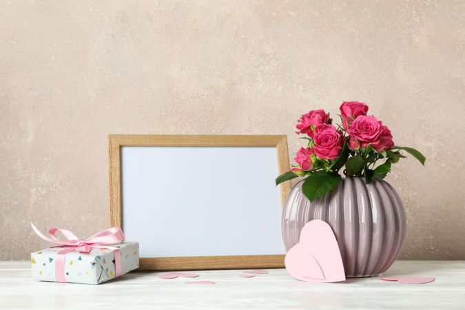 Vase with pink roses, empty frame, gift and little hearts on table against light brown background