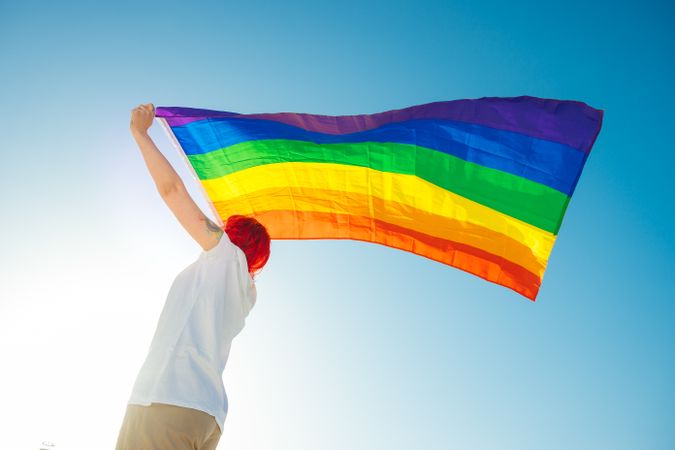 Young woman with red hair waving rainbow flag under blue sky