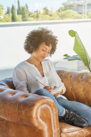 Serene Black Latina woman sitting in armchair and writing in diary