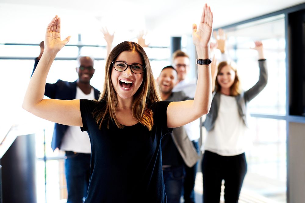 Businesswoman pictured in a bright office in front of her colleagues with their arms in the air