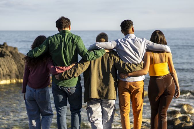Rear view of diverse group of friends embracing while looking at sea