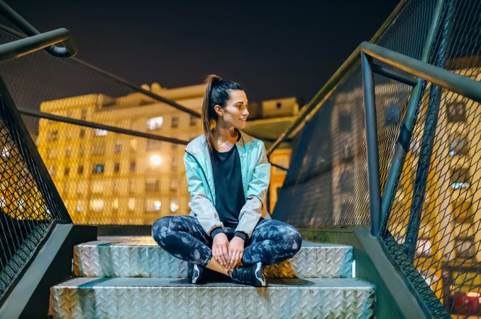 Happy female athlete wearing casual sportswear sitting on a metallic stairs at night