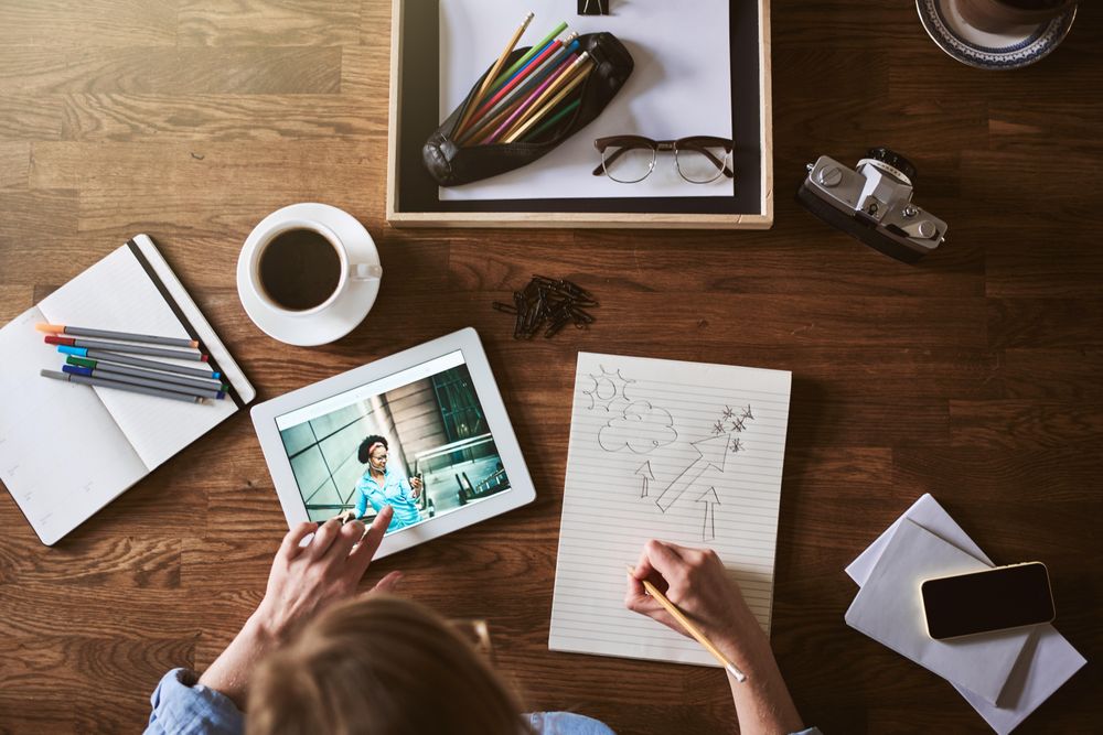 Top view of woman sketching with coffee, camera, and tablet on table