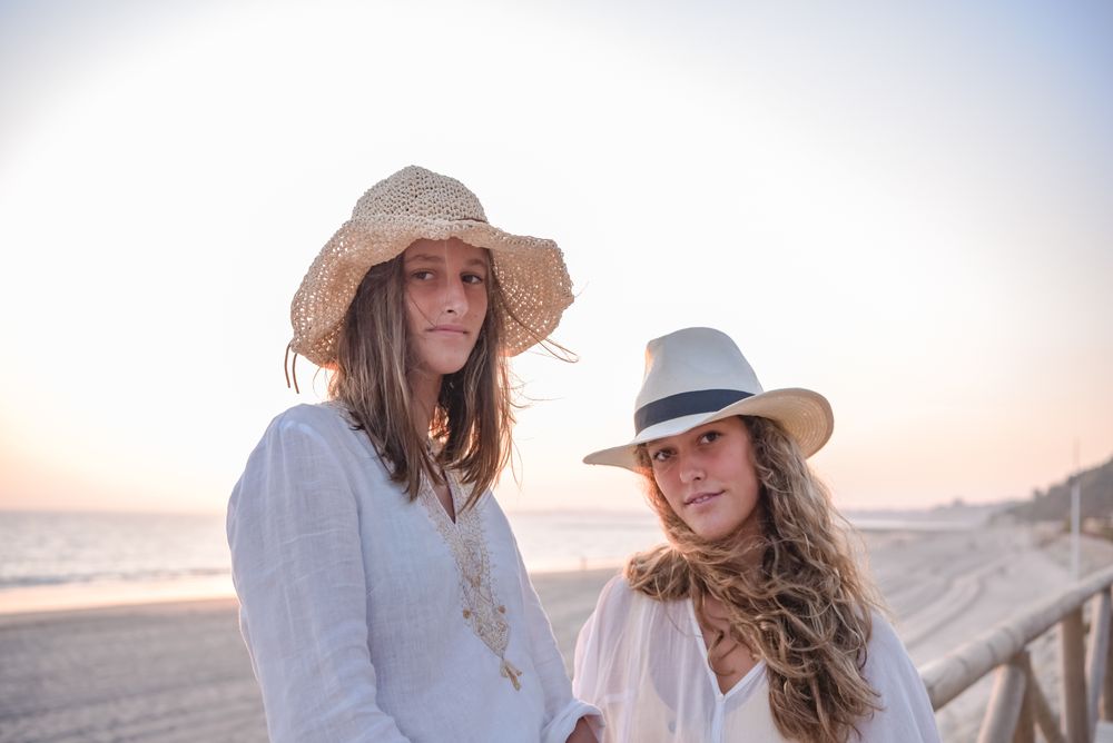 Smiling girlfriends in hats talking sitting on wooden fence on beach