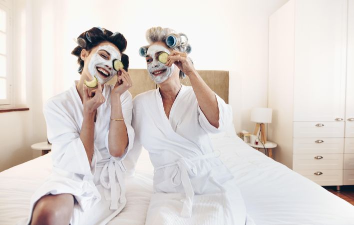 Smiling mother and daughter in bathrobes with face pack holding slices of cucumber to eyes