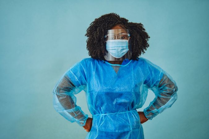 Black female doctor professional in surgical PPE, eye mask and shield, with hands on hip