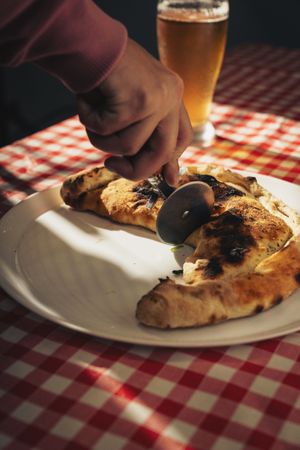 Calzone being sliced with pizza cutter on plate at checkered table with beer