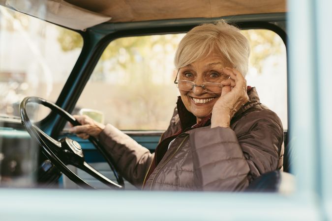 Woman sitting in driving seat of car holding her glasses down to look at the camera