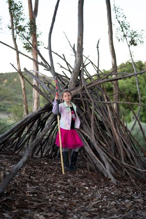 Confident young girl standing in front of tree fort holding a walking stick