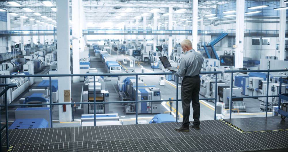 Experienced Male Engineer Standing on a Platform, Using Laptop Computer and Overlooking Production at a Modern Automated Electronics Manufacture with AI Support. Factory Executive at Work