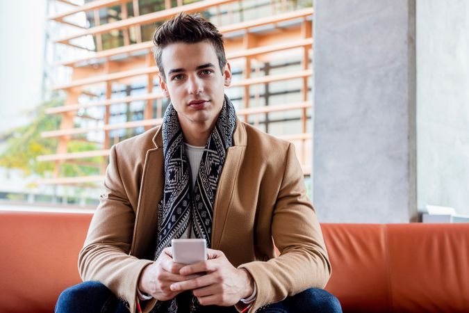 Adult man sitting outdoor on the bench in autumn holding mobile phone and looking at camera