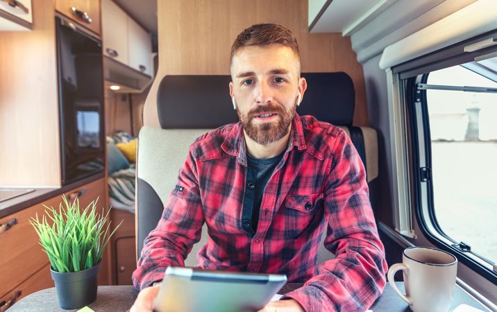 Man in red checkered shirt smiling sitting in van with digital tablet and coffee