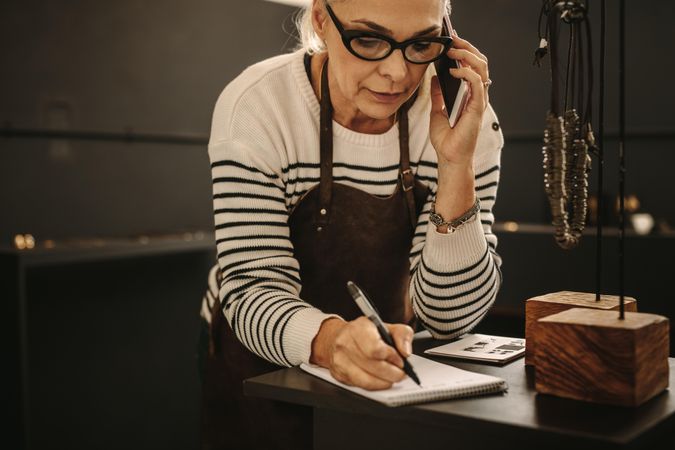 Portrait of mature female jeweler talking on mobile phone and making notes