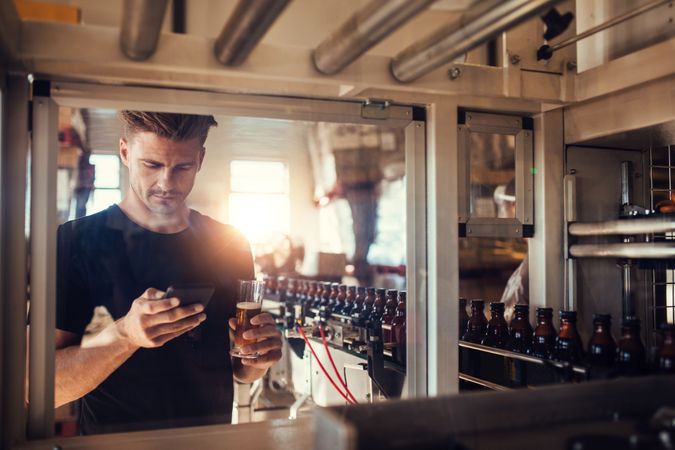 Young man with glass of beer at brewery plant using smart phone