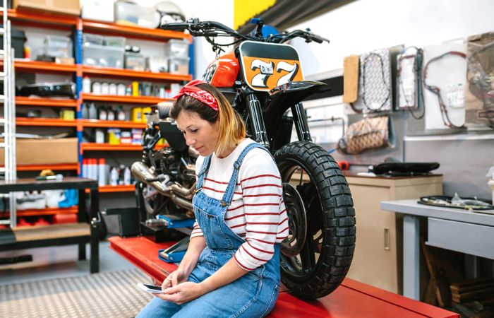 Mechanic woman looking phone sitting over platform with motorcycle on factory