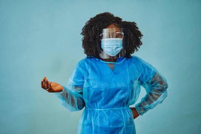 Black female doctor professional in surgical PPE, eye mask and shield, with hand on hip