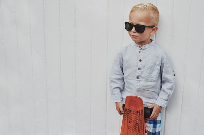 Young serious blond boy leaning against a wall with red skateboard, copy space