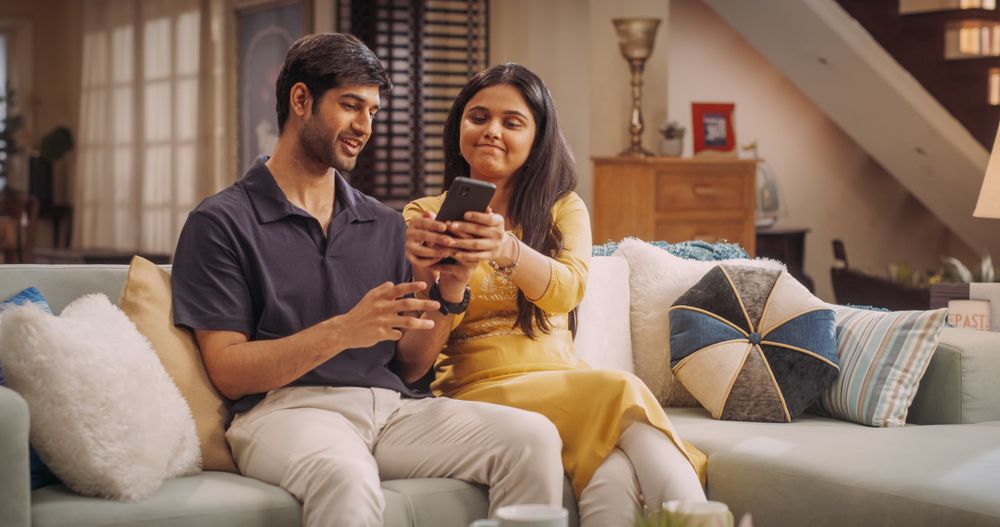 Young Indian Couple Sitting On The Sofa at Home: Woman Annoyed at her Boyfriend for Spending Too Much Time Online on Smartphone.