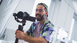 Smiling Young Male Videographer With Beard, Wearing Glasses And A Colorful Jacket, Operates A Professional Camera In A Well-lit Studio, Capturing Engaging Content For Film Or Advertising.