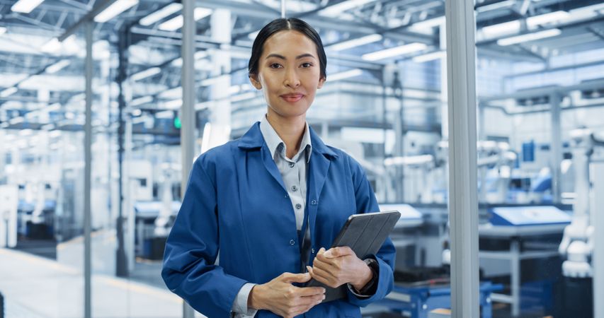 Portrait of a Young Asian Factory Technician Holding a Tablet Computer and Looking at Camera, Smiling. Industrial Female Specialist Wearing a Blue Coat, Working in a Modern Electronics Factory