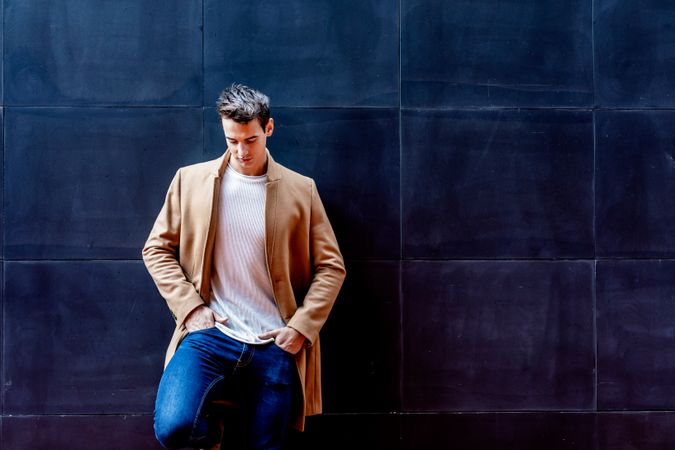 Cool male in camel coat looking down leaning against dark wall
