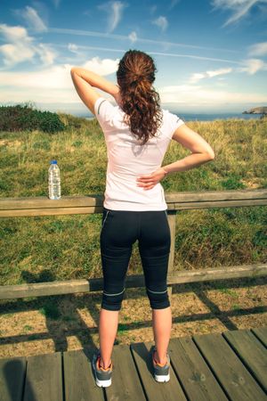 Woman in athletic gear holding hurt upper shoulders overlooking coast, vertical