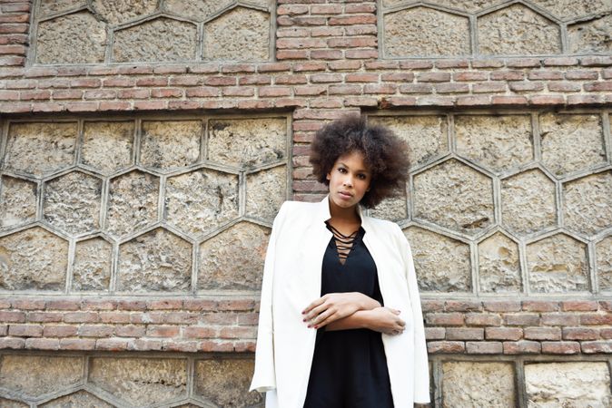 Female in dress and blazer leaning on brick wall and looking at camera
