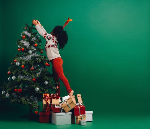 Young child standing on wrapped gifts reaching for the top of Christmas tree