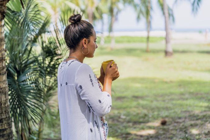 Relaxed woman enjoying hot drink and standing on exotic lawn