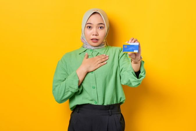 Muslim woman in headscarf and green blouse holding credit card with hand to her chest in surprise