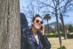 Side view of a young laughing hipster woman standing and leaning on a tree at park in sunny day while using a mobile phone