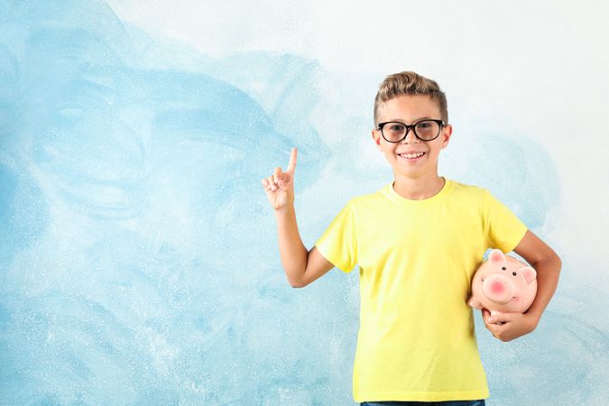Boy in glasses with piggy bank against color background, space for text