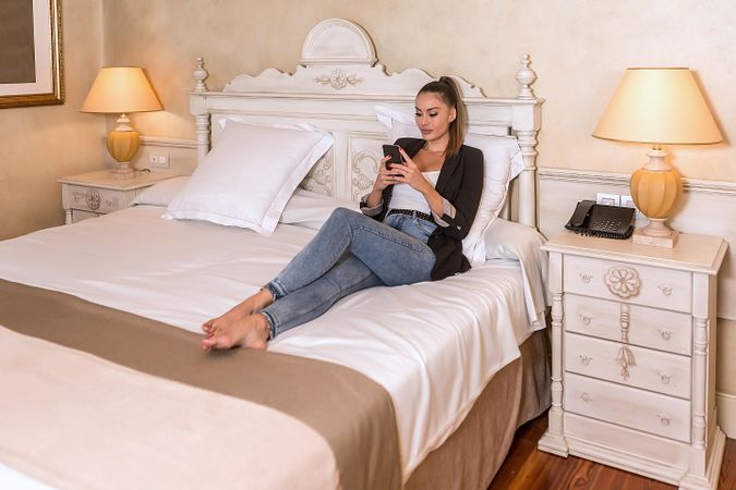 Woman in dark blazer using smartphone and sitting on bed