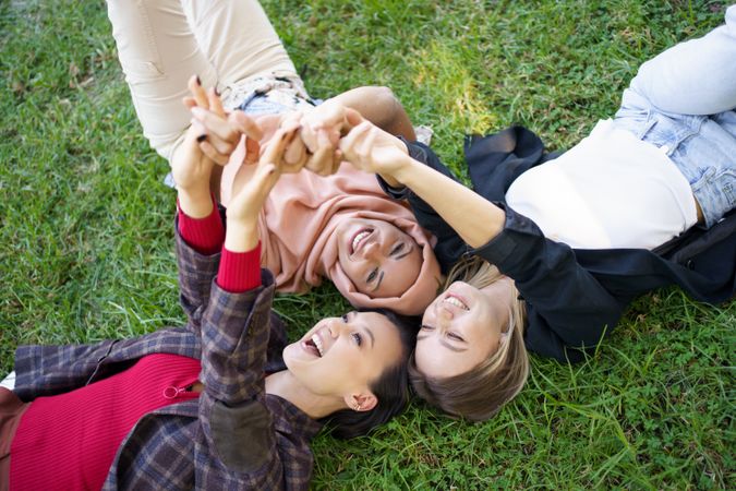 Looking down at three happy friends lying on grass with their arms joined above their heads