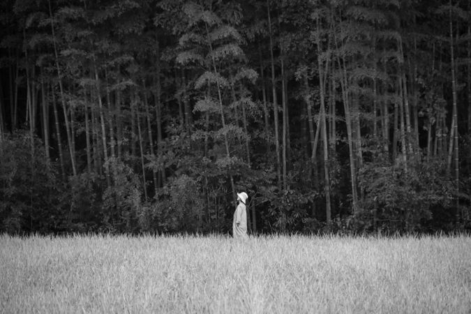 Grayscale photo of a person with a hat standing on grass near woods