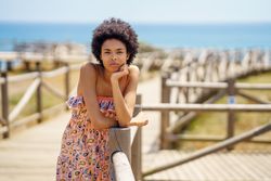 Serious woman with afro hair standing on wooden footpath by the coast leaning on hand rail
