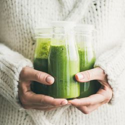 Woman holding three bottles of green smoothie wearing cozy sweater, square crop