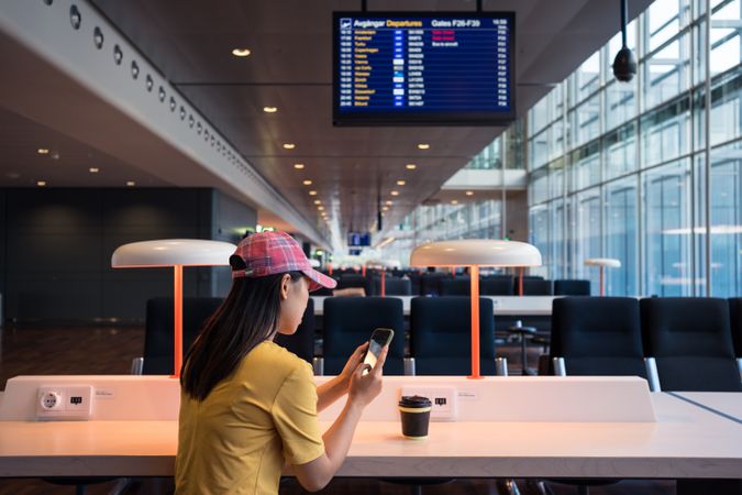 Asian woman browsing smartphone while drinking coffee at table under lighting lamp