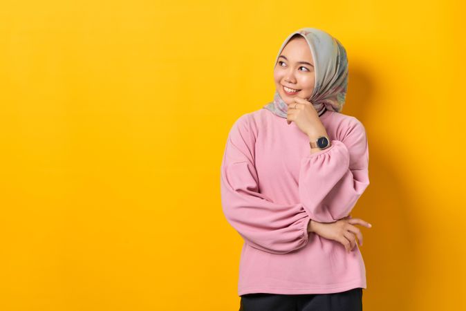 Smiling Muslim woman in headscarf holding smart phone and looking away in thought with hand on chin