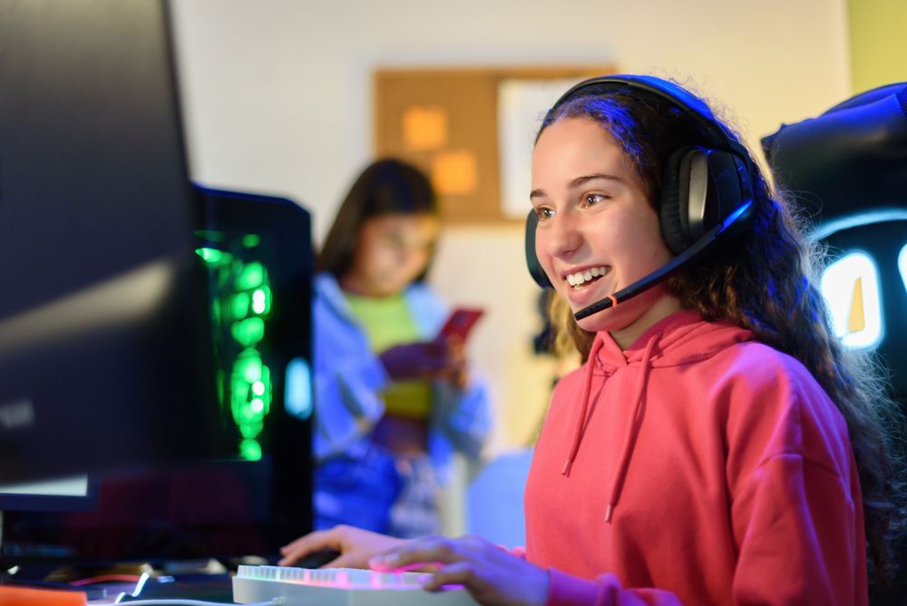 Happy teenage girl in headphones sitting at table and playing games in room