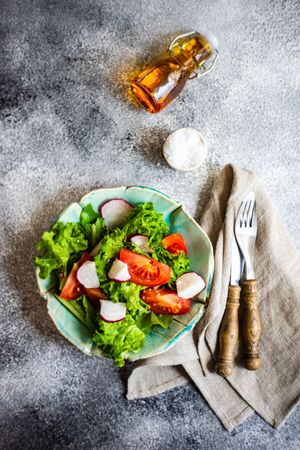 Bowl of salad with tomatoes, radish & lettuce on marble counter with oil & salt