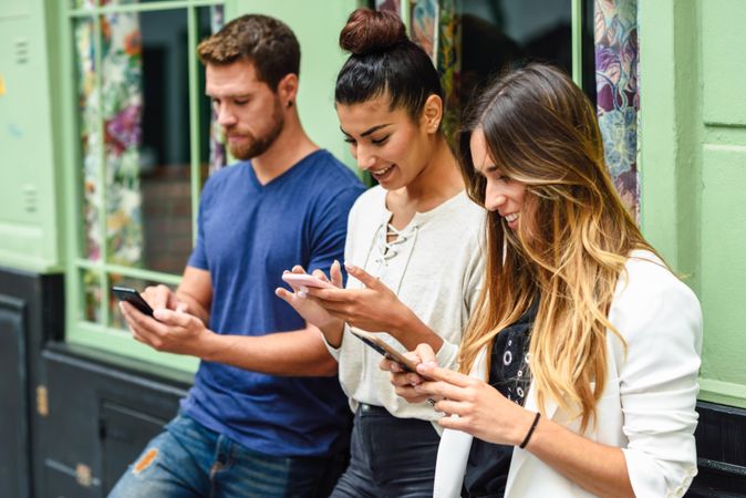 Two female and one male friend looking down at their phones outside