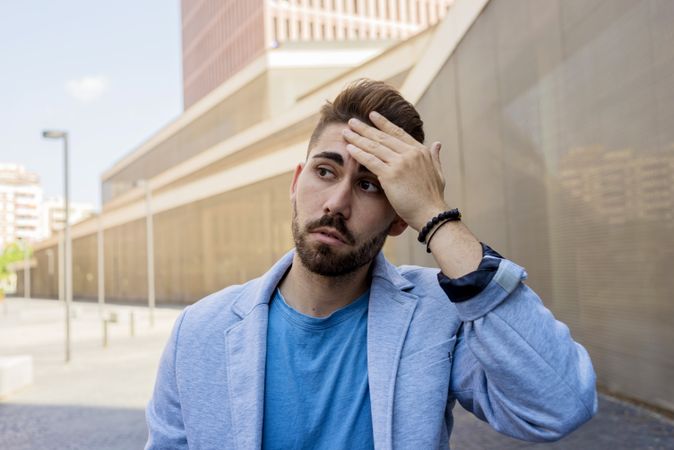 Young bearded man in front of building wearing smart casual with hand to his forehead