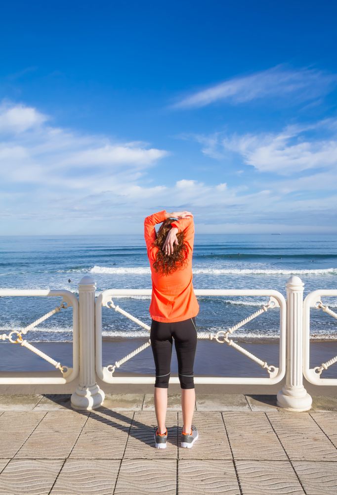 Athletic woman stretching triceps before running on the beach promenade on a summer morning, vertical