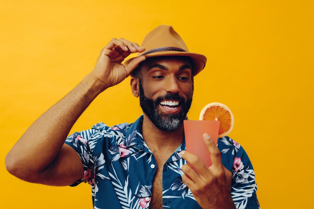 Content male adjusting hat while wearing colorful shirt and holding cocktail with yellow background
