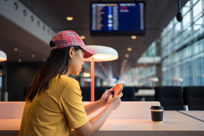 Asian woman browsing smartphone while drinking coffee at table under lighting lamp